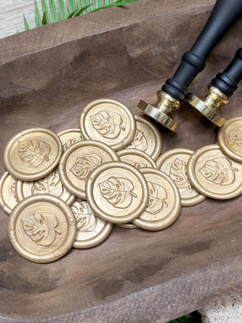 May include: Gold wax seals with a monstera leaf design are scattered in a wooden bowl. Two black and gold wax seal stamps are also in the image. The seals are round and have a raised leaf pattern. The bowl is a warm brown color.