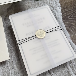 May include: A white wedding invitation with a black border and a wax seal with a floral design. The invitation is tied with black ribbon and is on a white fabric background.