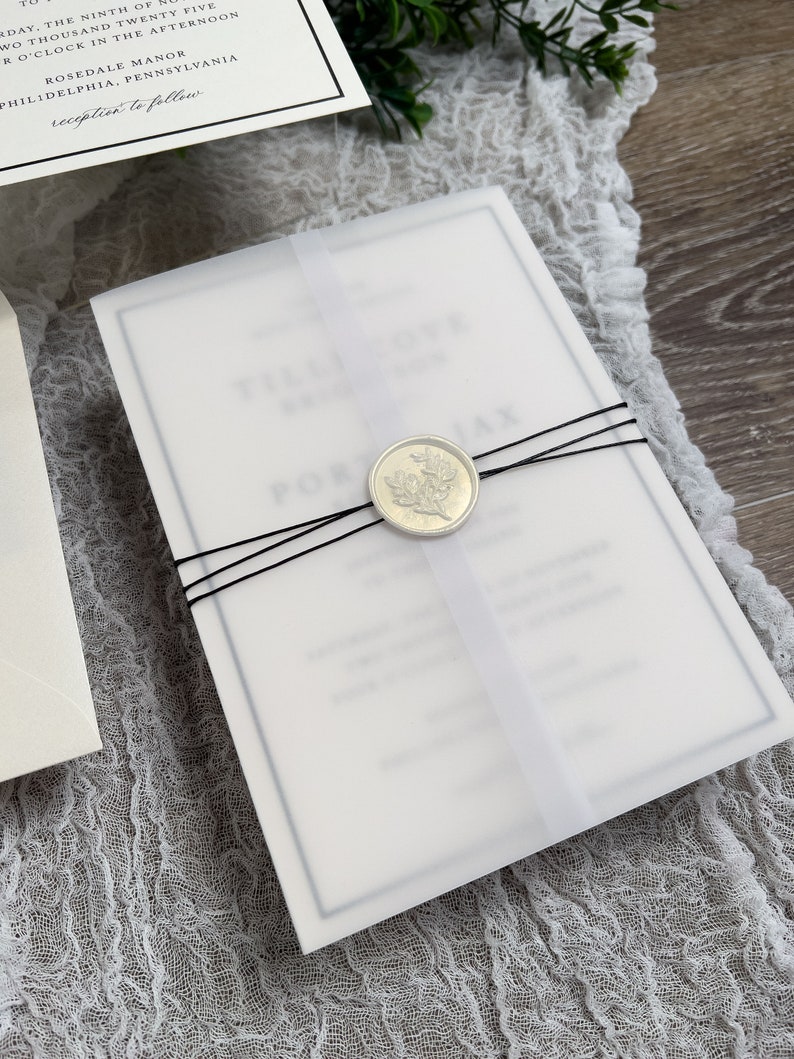 May include: A white wedding invitation with a black border and a wax seal with a floral design. The invitation is tied with black ribbon and is on a white fabric background.