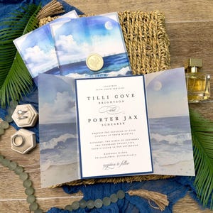 May include: Wedding invitation suite featuring a watercolor ocean scene with a full moon. The invitation includes the names Tilli Cove Brightson and Porter Jax Schearer, along with the wedding date and location. The suite is accented with a gold wax seal and a blue fabric.
