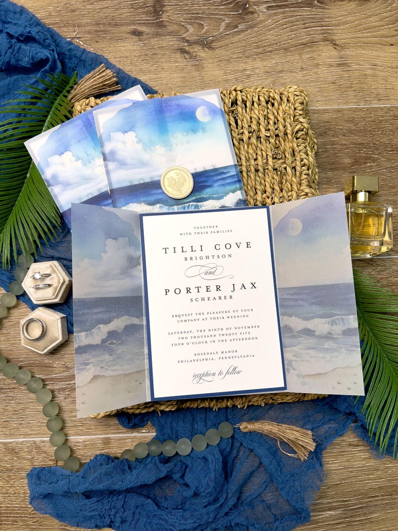 May include: Wedding invitation suite featuring a watercolor ocean scene with a full moon. The invitation includes the names Tilli Cove Brightson and Porter Jax Schearer, along with the wedding date and location. The suite is accented with a gold wax seal and a blue fabric.