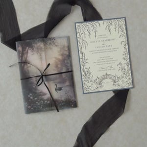 Cloud Ivory wedding invitation featuring delicate botanical border illustrations displayed beside a translucent woodland lake vellum jacket tied with black ribbon, creating a dreamy Swan Lake-inspired wedding stationery design.