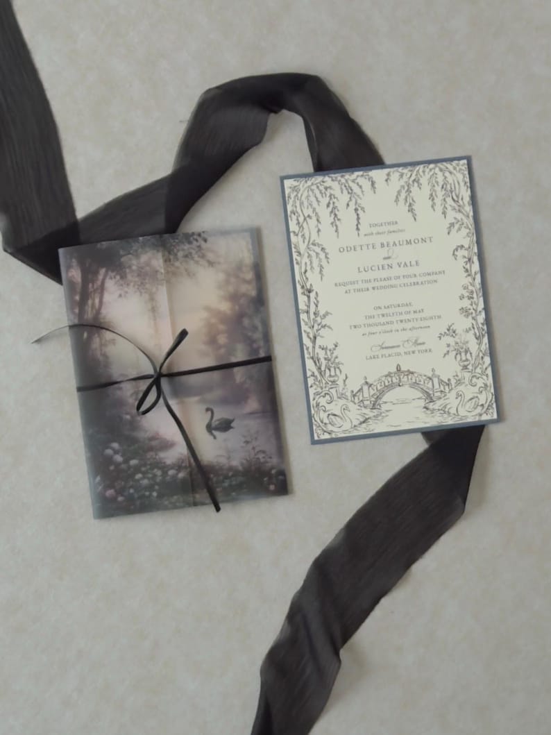 Cloud Ivory wedding invitation featuring delicate botanical border illustrations displayed beside a translucent woodland lake vellum jacket tied with black ribbon, creating a dreamy Swan Lake-inspired wedding stationery design.
