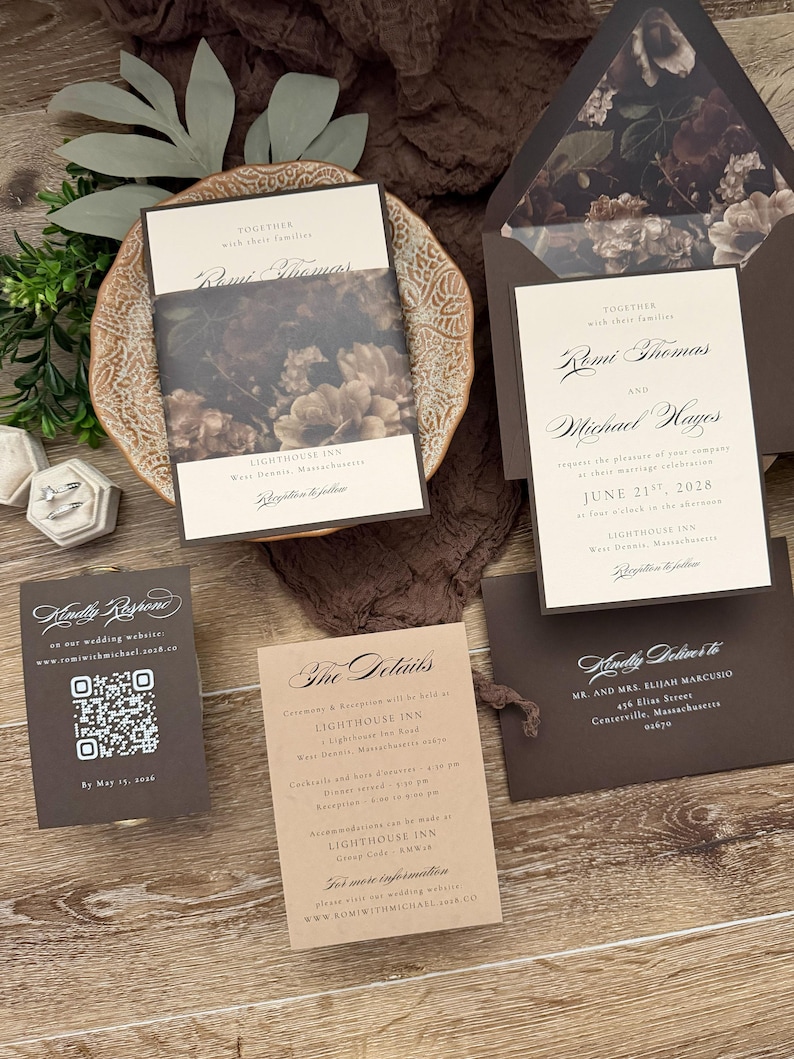 May include: A wedding invitation suite featuring cream-colored cards with dark brown text, a dark floral envelope liner, and a matching belly band. Includes a QR code response card, a details card, and an addressed envelope with dark brown accents.