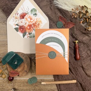 Puede incluir: Una invitaci&oacute;n de boda con un sobre naranja &oacute;xido y un sobre interior blanco con un dise&ntilde;o floral. La invitaci&oacute;n est&aacute; atada con cordel y sellada con un sello de cera verde. El texto de la invitaci&oacute;n dice "&Uacute;nete a nosotros para la boda de" y "Todos los detalles responde amablemente".