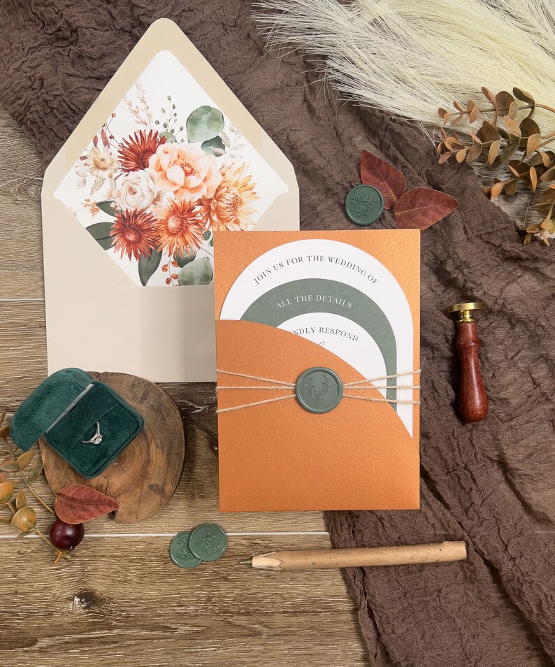 Puede incluir: Una invitaci&oacute;n de boda con un sobre naranja &oacute;xido y un sobre interior blanco con un dise&ntilde;o floral. La invitaci&oacute;n est&aacute; atada con cordel y sellada con un sello de cera verde. El texto de la invitaci&oacute;n dice "&Uacute;nete a nosotros para la boda de" y "Todos los detalles responde amablemente".