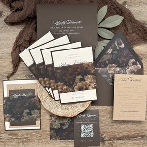 May include: A wedding invitation suite featuring dark brown envelopes, cream-colored cards, and belly bands with a dark floral print. The set includes a mailing envelope addressed to Ms. Katelyn Greene, a details card, and a response card with a QR code.