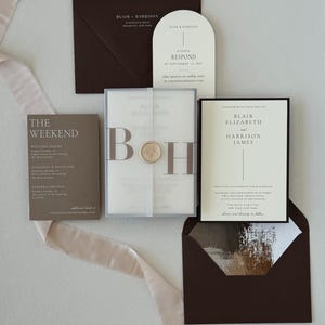 Modern neutral wedding stationery featuring a brown details card, ivory arch RSVP card, bordered invitation and translucent monogram vellum wrap styled with soft ribbon.