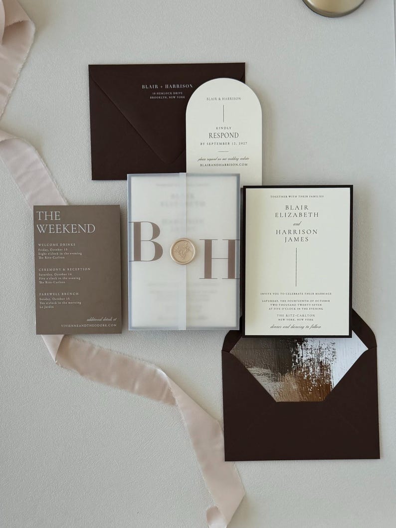 Modern neutral wedding stationery featuring a brown details card, ivory arch RSVP card, bordered invitation and translucent monogram vellum wrap styled with soft ribbon.