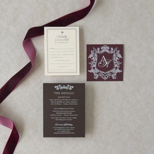 May include: A set of three wedding stationery cards in cream and deep burgundy. One card features a burgundy crest with the initials AH, another is a cream RSVP card, and the third is a burgundy details card. A long burgundy velvet ribbon is included.