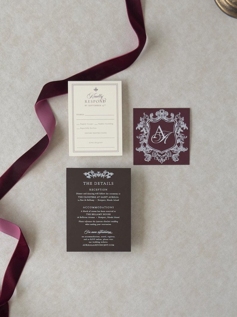May include: A set of three wedding stationery cards in cream and deep burgundy. One card features a burgundy crest with the initials AH, another is a cream RSVP card, and the third is a burgundy details card. A long burgundy velvet ribbon is included.