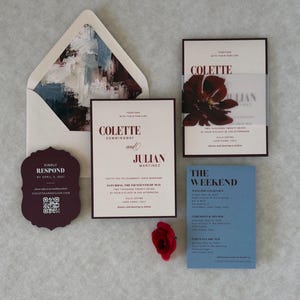 Burgundy and dusty blue floral wedding invitation suite featuring an ivory invitation with dark floral artwork, dusty blue details card, burgundy scalloped RSVP card, and burgundy mailing envelope with coordinating floral liner.