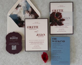 May include: A wedding invitation suite featuring cream-coloured cards with dark red text, a dark red response card with a QR code, a blue weekend itinerary card, and an envelope with an abstract blue, white, and dark red painted liner design.