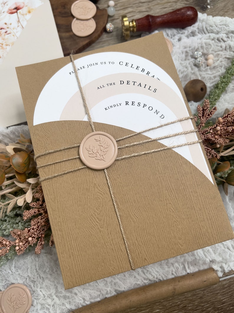 May include: A brown invitation card with a white and pink layered design. The card is tied with twine and sealed with a wax stamp. The text on the card reads "Please join us to celebrate... all the details... kindly respond".
