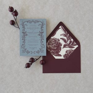 Dusty blue wedding invitation displayed with burgundy A7 envelope and vintage botanical floral envelope liner