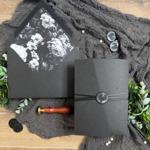 Puede incluir: Invitaci&oacute;n de boda negra con un dise&ntilde;o floral en el sobre interior y un sello de cera negro en el sobre exterior.