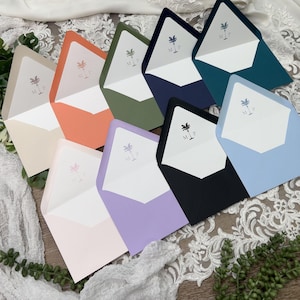 May include: A collection of colorful envelopes in various shades, including peach, green, and blue. Each envelope features a white interior with a palm tree and initials. The envelopes are arranged on a wooden surface with white lace and greenery.