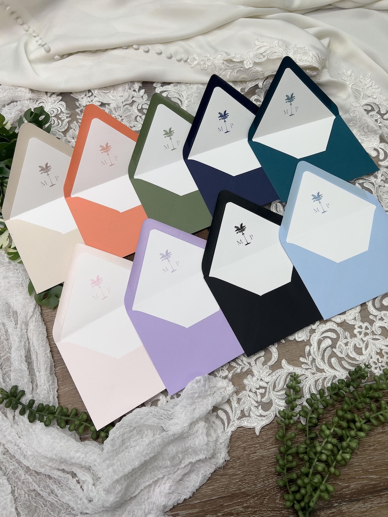 May include: A collection of colorful envelopes in various shades, including peach, green, and blue. Each envelope features a white interior with a palm tree and initials. The envelopes are arranged on a wooden surface with white lace and greenery.