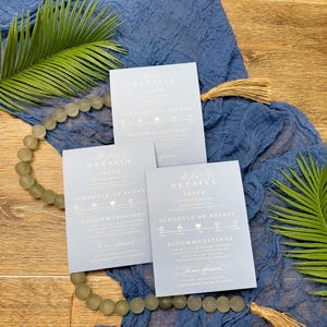 May include: Three light blue wedding invitation cards with the text "The Fine Details" and event information. The cards are arranged on a blue fabric with a beaded garland and palm leaves.