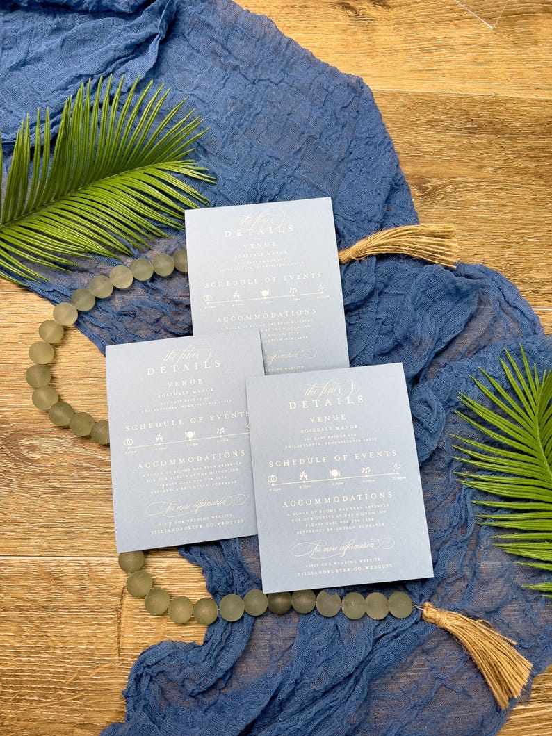 May include: Three light blue wedding invitation cards with the text "The Fine Details" and event information. The cards are arranged on a blue fabric with a beaded garland and palm leaves.