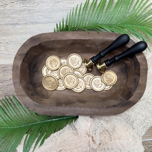 May include: A wooden bowl filled with gold wax seals and two black-handled wax stampers. The seals have a leaf design. The bowl sits on a wooden surface with green fern leaves and a beige cloth.