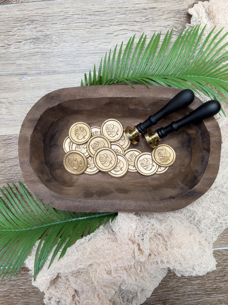 May include: A wooden bowl filled with gold wax seals and two black-handled wax stampers. The seals have a leaf design. The bowl sits on a wooden surface with green fern leaves and a beige cloth.