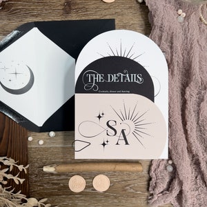 May include: A wedding invitation with a black and white design featuring a crescent moon and stars on the left side and a sunburst with the text "THE DETAILS" on the right side. The text "Cocktails, dinner and dancing" is below the sunburst. The invitation is on a wooden surface with a pink fabric and wax seals.