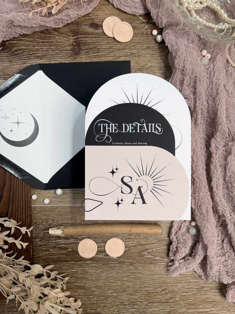 May include: A wedding invitation with a black and white design featuring a crescent moon and stars on the left side and a sunburst with the text "THE DETAILS" on the right side. The text "Cocktails, dinner and dancing" is below the sunburst. The invitation is on a wooden surface with a pink fabric and wax seals.