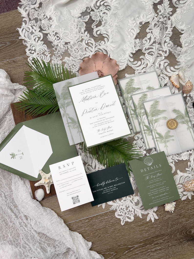 May include: An assortment of wedding stationery featuring a tropical theme. The invitations, RSVP cards, and details cards are in shades of green and white, with palm tree illustrations. The stationery is arranged with seashells and greenery.