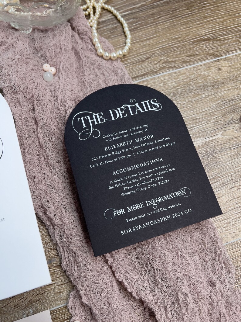 May include: Black and white wedding invitation with a decorative arch design. The invitation details the ceremony and reception location, time, and contact information for accommodations. The text reads "The Details", "Elizabeth Manor", "223 Eastern Ridge Street, New Orleans, Louisiana", "Cocktail Hour at 5:00 pm | Dinner served at 6:00 pm", "Accommodations", "A block of rooms has been reserved at The Hilton Garden Inn with a special rate", "Please call 800.233.1234", "Wedding Group Code: V12024", "For More Information", "Please visit our wedding website: SORAYAANDASPEN.2024.CO".