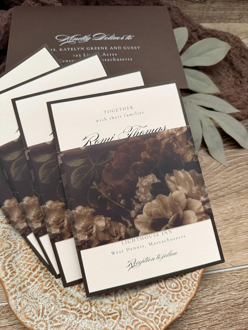 May include: A stack of cream-colored wedding invitations featuring dark brown borders and a floral belly band with dark brown and beige roses. The text reads Together with their families, Romi Thomas, Lighthouse Inn, West Dennis, Massachusetts, and Reception to follow.