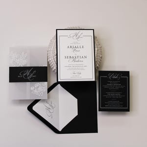 Puede incluir: Un conjunto de invitaciones de boda que incluye una tarjeta blanca con texto negro, un sobre negro con forro blanco, una funda de papel vegetal con flores blancas y una faja negra, adem&aacute;s de una tarjeta de detalles en color negro.