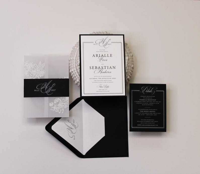 Puede incluir: Un conjunto de invitaciones de boda que incluye una tarjeta blanca con texto negro, un sobre negro con forro blanco, una funda de papel vegetal con flores blancas y una faja negra, adem&aacute;s de una tarjeta de detalles en color negro.