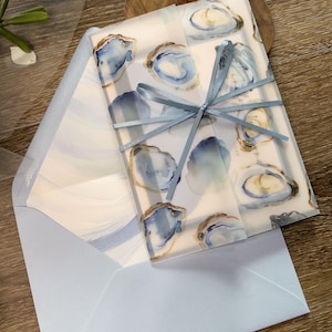 May include: Stationery set with a watercolour oyster design in blue and gold. The stationery is tied with a light blue ribbon and includes a matching light blue envelope. The design is elegant and suitable for invitations or announcements.