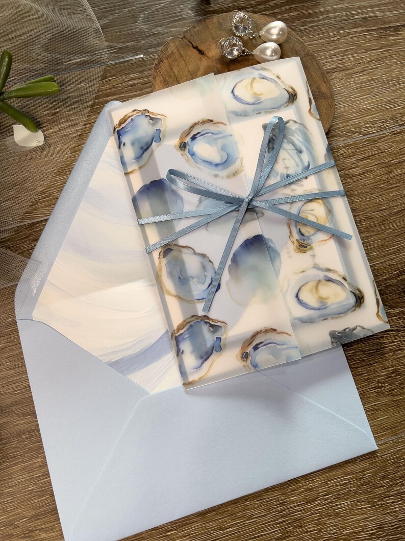 May include: Stationery set with a watercolour oyster design in blue and gold. The stationery is tied with a light blue ribbon and includes a matching light blue envelope. The design is elegant and suitable for invitations or announcements.