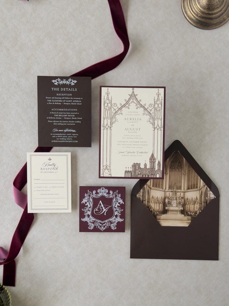 Aurelia Gothic castle wedding invitation flat lay showing architectural vellum overlay, cream invitation, Bitter Chocolate details card and envelope, Cashmere RSVP envelope, and ornate Claret tag on a light textured background.