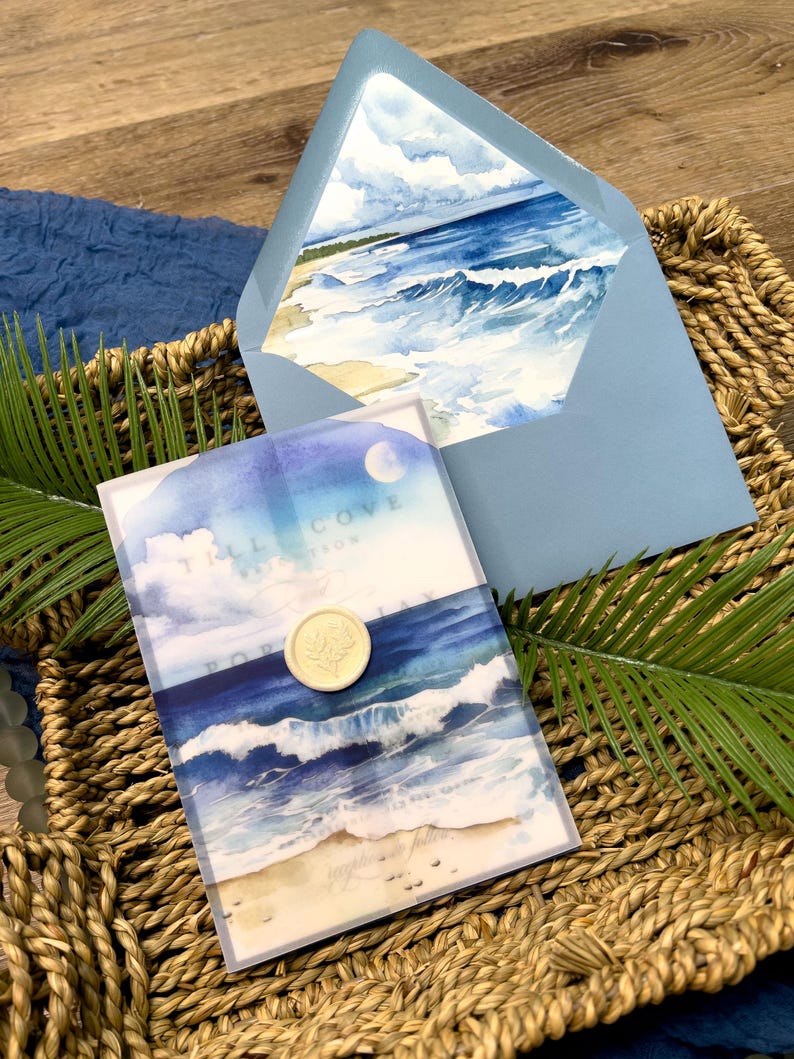 May include: A light blue envelope and invitation featuring a watercolor painting of ocean waves and a sandy beach. The invitation is sealed with a gold wax seal. The text "Tall Cove" is visible on the invitation.