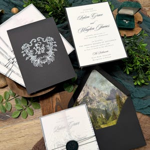 May include: Elegant wedding invitation suite featuring a black invitation with a floral monogram, a white invitation with calligraphy, and a clear invitation with a mountain design. The suite includes a black envelope with a mountain landscape and a ring in a green box.