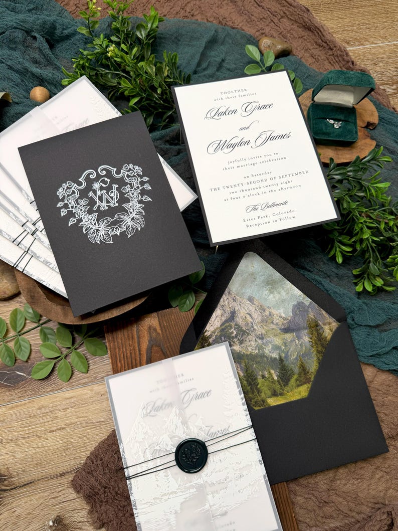 May include: Elegant wedding invitation suite featuring a black invitation with a floral monogram, a white invitation with calligraphy, and a clear invitation with a mountain design. The suite includes a black envelope with a mountain landscape and a ring in a green box.