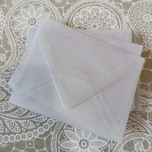 May include: A stack of translucent white envelopes. The envelopes are square with a flap closure. The background is a patterned fabric with a beige and white floral design.