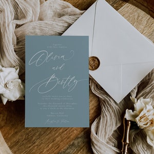 May include: A wedding invitation suite featuring a dusty blue invitation card with elegant script, an envelope with a gold wax seal, and decorative elements. The invitation reads "Olivia and Bentley" and includes event details.
