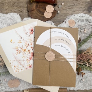 May include: A brown invitation with a white circle and the text "Please join us to celebrate... all the details... kindly respond". The invitation is tied with twine and sealed with a wax stamp. The invitation is on a wooden surface with a white lace overlay.