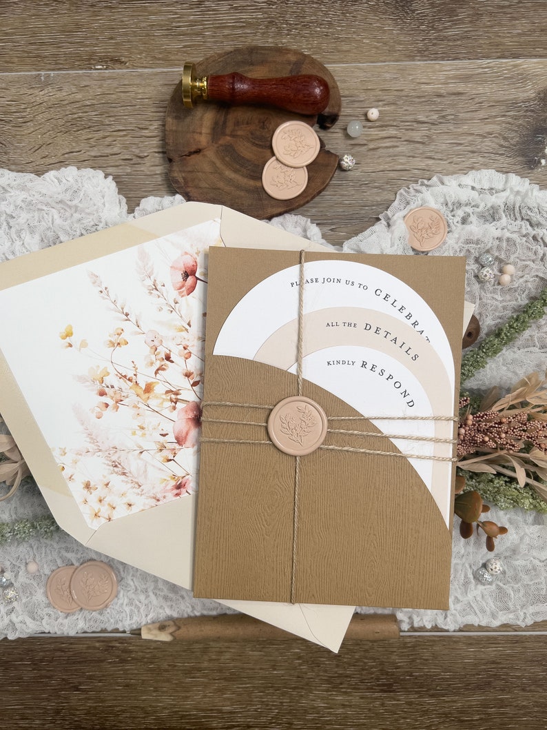 May include: A brown invitation with a white circle and the text "Please join us to celebrate... all the details... kindly respond". The invitation is tied with twine and sealed with a wax stamp. The invitation is on a wooden surface with a white lace overlay.
