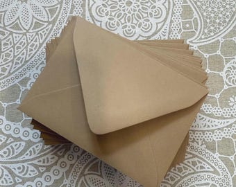 May include: A stack of kraft paper envelopes, in a warm brown hue, arranged on a patterned fabric. The envelopes are triangular in shape, with a folded flap. The fabric background features a repeating white floral design.