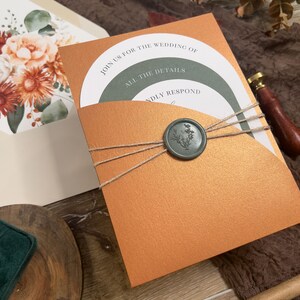 Puede incluir: Una invitaci&oacute;n de boda de color cobre con un bolsillo interior verde y blanco. La invitaci&oacute;n tiene un sello de cera con un dise&ntilde;o floral y el texto "Join us for the wedding of" y "All the details" en el frente.