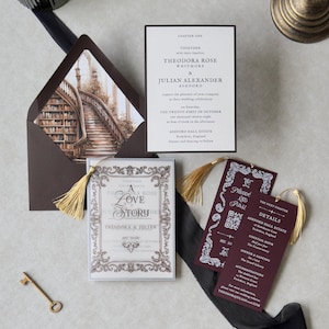 Vintage-inspired Theodora invitation suite with ornate ivory stationery, burgundy details card, decorative bookmark, and dark brown envelope with library staircase liner, arranged with antique brass styling accents.