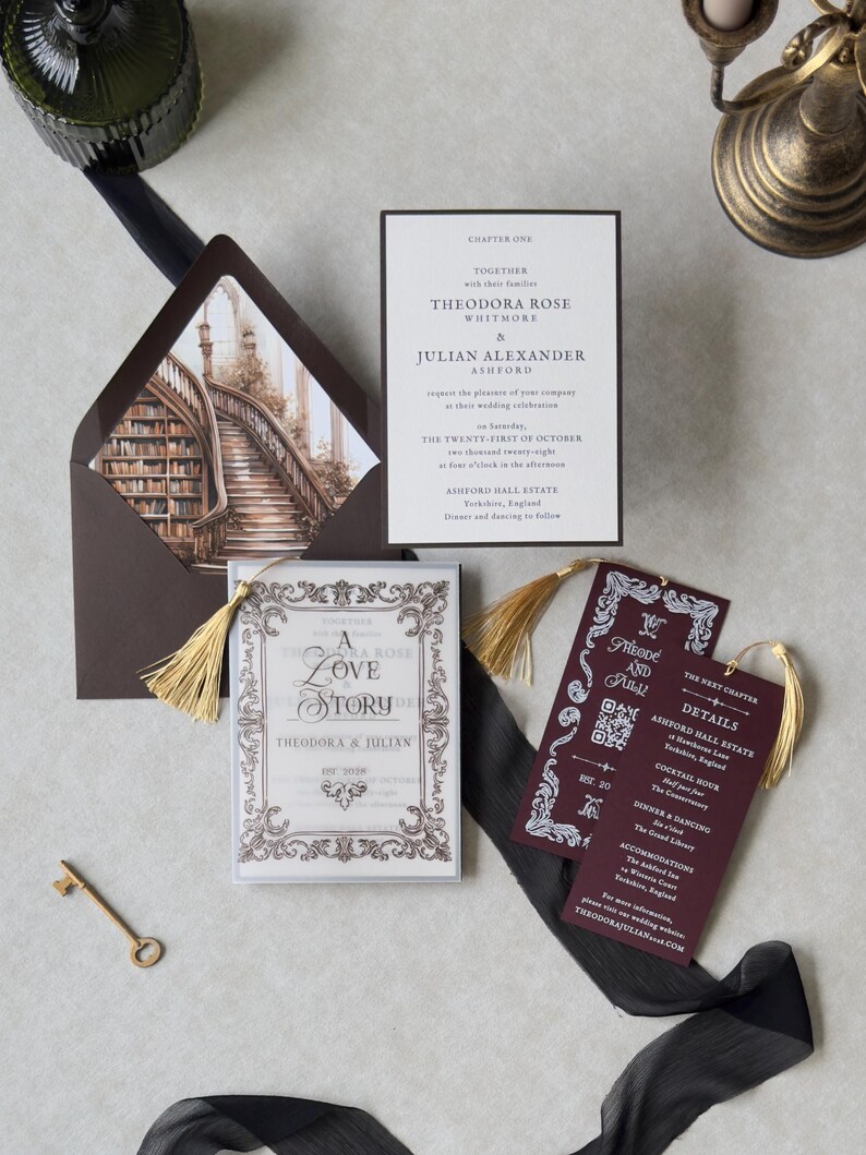Vintage-inspired Theodora invitation suite with ornate ivory stationery, burgundy details card, decorative bookmark, and dark brown envelope with library staircase liner, arranged with antique brass styling accents.
