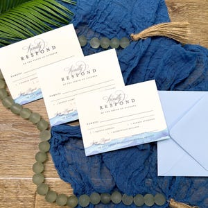 May include: Wedding response cards with blue watercolor accents and the text "Kindly Respond by the Tenth of October." The cards are on a blue fabric with a light blue envelope and decorative beads.