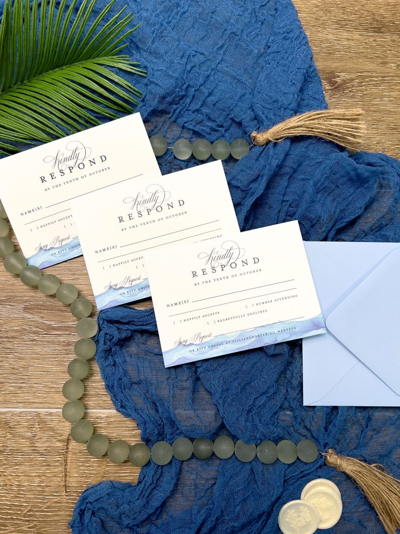 May include: Wedding response cards with blue watercolor accents and the text "Kindly Respond by the Tenth of October." The cards are on a blue fabric with a light blue envelope and decorative beads.