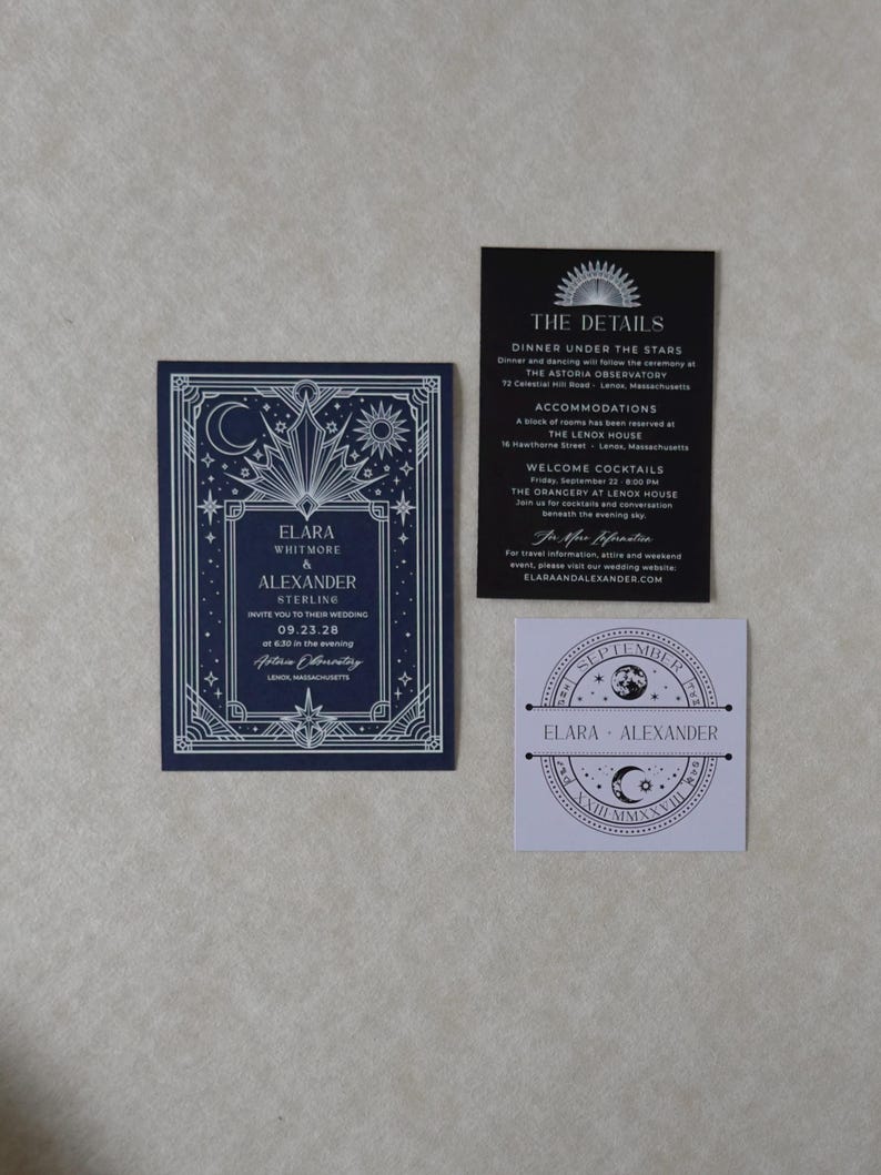 Elara celestial wedding stationery collection arranged in a flat lay, showing the navy invitation with silver geometric sun and moon border, black details card with white ink, and lavender celestial tag.